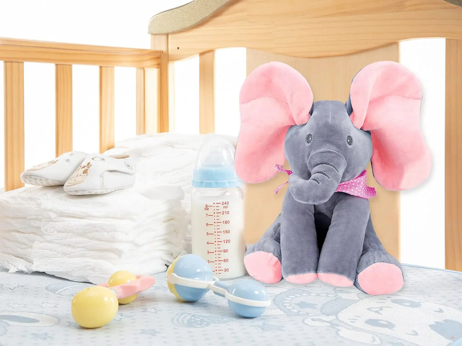 Un jouet éléphant en peluche doux BABY-PREMA est posé sur la table à langer d'un bébé prématuré, entouré d'une paire de chaussures pour bébé, d'un biberon et de hochets, créant un cadre de chambre de bébé confortable et nourrissant.