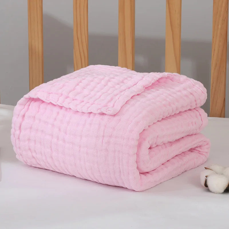 Une couverture Couvertures Mousseline rose tendre soigneusement pliée sur une surface blanche avec un fond en bois, dégageant une ambiance douillette et chaleureuse, parfaite comme accessoire pour bébé de BABY PREMA.