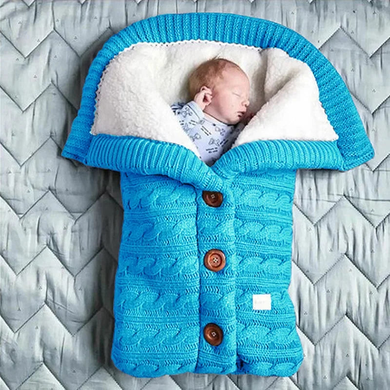 Un nouveau-né dort paisiblement emmailloté dans une couverture de poussette pour bébé BABY PREMA en tricot bleu douillet en forme de gigoteuse, avec des boutons en bois ajoutant une touche de charme.