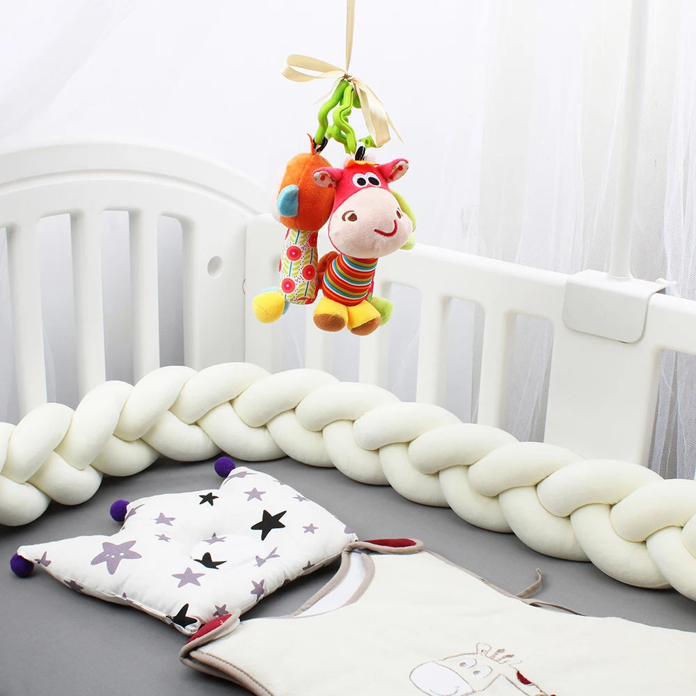 Un lit de bébé blanc avec un tour de lit doux et tressé sur les côtés, des jouets en peluche colorés suspendus et un oreiller blanc orné d'étoiles violettes et noires reposent sur un matelas gris. Une gigoteuse blanche ornée d'un personnage de dessin animé est également nichée dans la Tour de lit bébé 4M tressé de BABY PREMA.