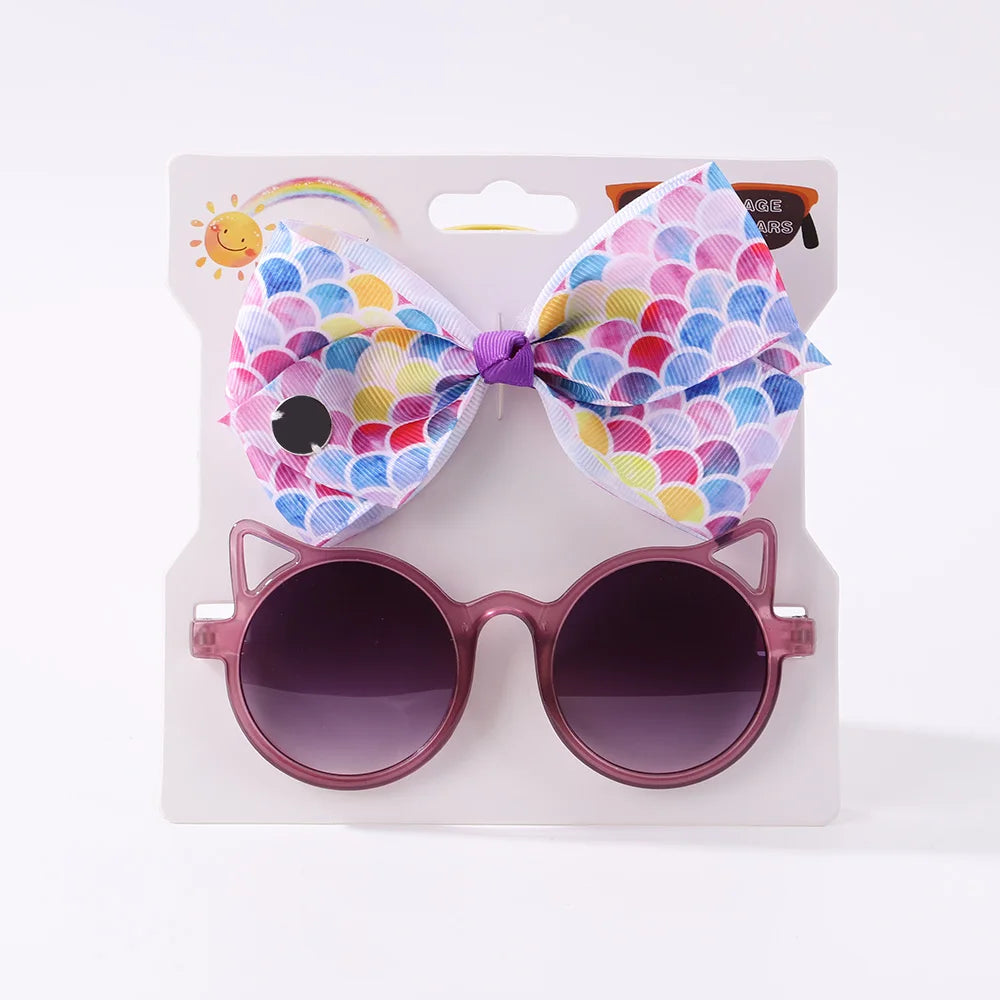 Une paire de lunettes de soleil violettes avec des montures d'oreilles de chat et un nœud coloré à motif d'écailles de poisson sont présentées sur du papier cartonné emballé, parfait comme Set d'Accessoires Bébé | Lunettes de Cheveux pour Bébé de BABY PREMA.