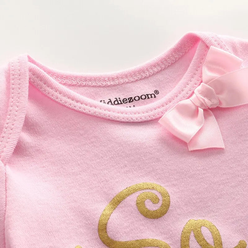 Gros plan d'un vêtement pour bébé rose avec le nom de la marque "BABY PREMA" sur l'étiquette du col, avec des manches courtes et un petit nœud ainsi qu'une lettrage doré partiel sur le devant. Le produit présenté fait partie du "Lot de 5 Pièces 100% Coton pour Bébé".