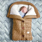 Un nouveau-né endormi dans une douillette couverture de poussette pour bébé BABY PREMA en forme de sac de couchage, ornée de boutons décoratifs et des « nécessaires pour bébé » brodés dessus, sur un fond texturé.