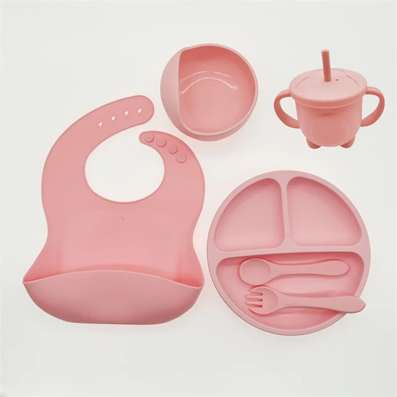 Ensemble d'alimentation pour bébé en silicone rose comprenant un bavoir, une assiette sectionnée, un bol, une tasse avec poignées et une paille, une fourchette et une cuillère disposées sur un fond blanc. Fabriqué à partir de silicone de haute qualité, cet Ensemble 6-8 Pièces de Vaisselle pour enfants de BABY PREMA assure durabilité et sécurité pour les repas de votre tout-petit.