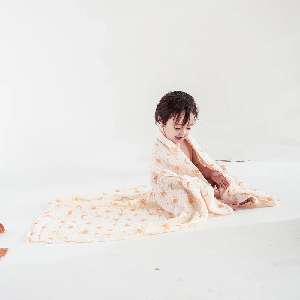 Une personne assise par terre enveloppée dans une délicate couverture Lange d'Emmaillotage aux motifs de BABY PREMA, apparaissant contemplative et sereine sur un fond blanc, tenant un bébé poids léger qui semble être en dodo.