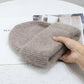 Une main tenant un bonnet tricoté beige plié et confortable Angora Pour Maman pour bébé de BABY PREMA sur un magazine qui repose sur une surface blanche texturée, suggérant une ambiance calme et élégante.