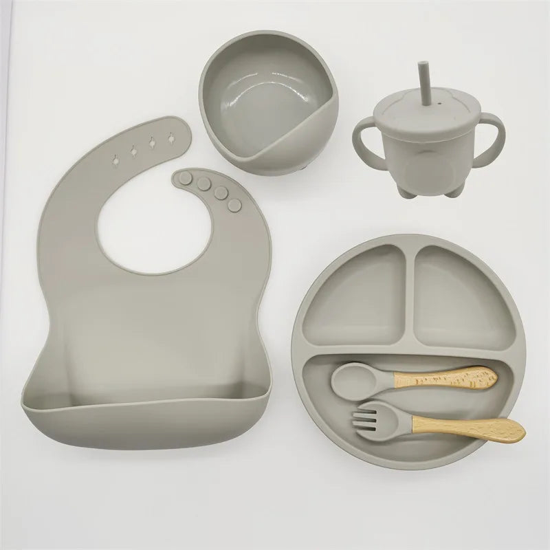 Un Ensemble 6-8 Pièces de Vaisselle pour enfants de BABY PREMA comprend un bavoir, un bol, un gobelet, une assiette divisée, une cuillère et une fourchette, le tout fabriqué en silicone de haute qualité de couleur gris clair.