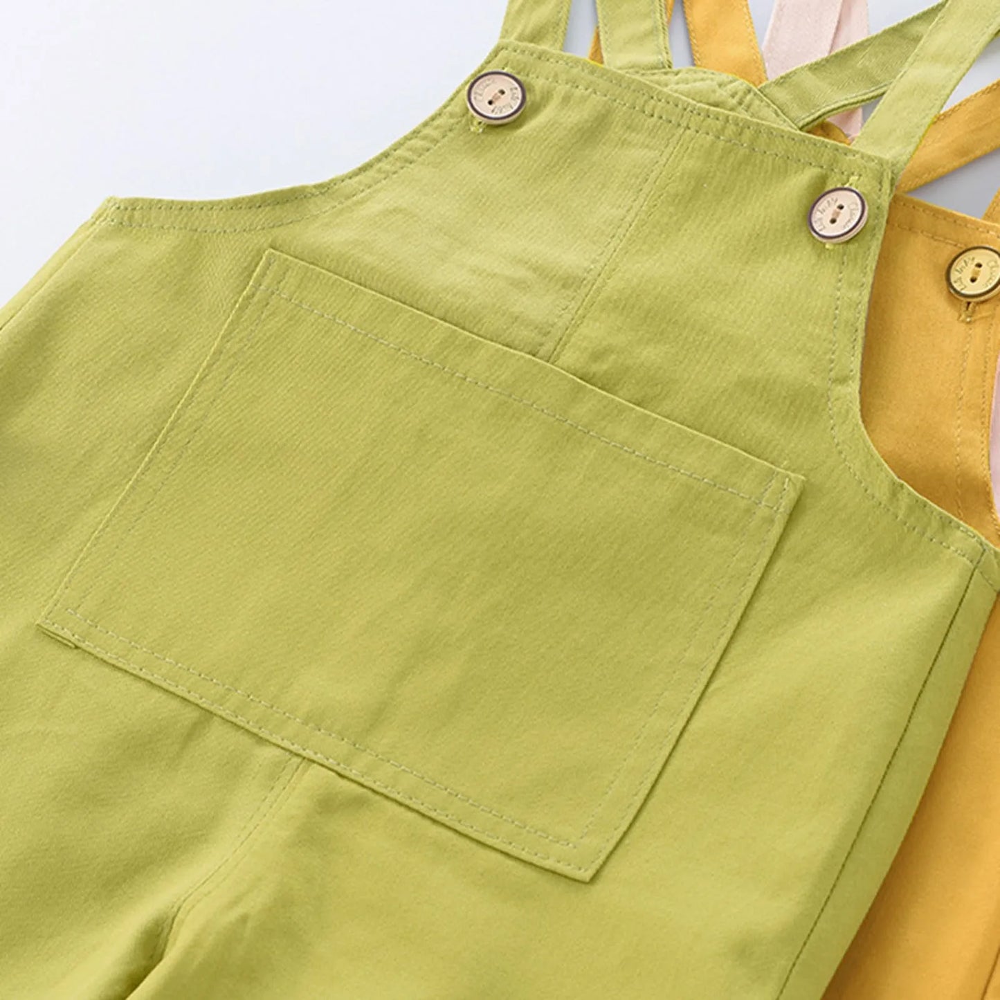 Gros plan de deux salopettes pour enfants sur une surface blanche, comportant des bretelles boutonnées et de grandes poches avant. L’un est vert et l’autre jaune. Ces adorables Salopette Bébé Unie de BABY-PREMA sont confectionnées en coton ou denim doux, assurant le confort de vos petits.