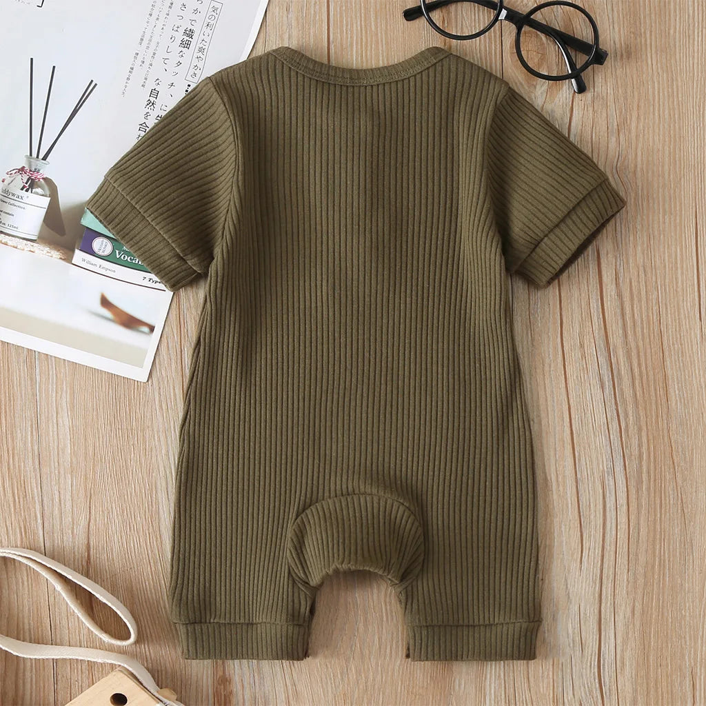 Une Barboteuse Bébé Manches Courtes vert olive côtelée de BABY-PREMA est disposée sur une surface en bois à côté d'un journal plié, de lunettes noires et d'une sangle beige. C'est le vêtement idéal pour les petits.