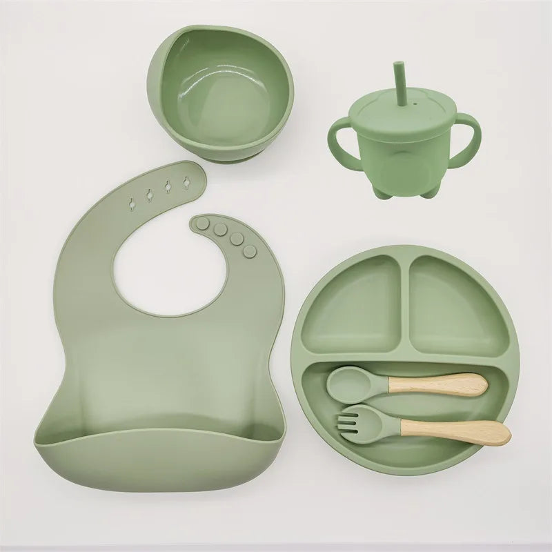 Un ensemble d'articles d'alimentation pour bébé verts en silicone de haute qualité, comprenant un bol, un gobelet avec poignées, une assiette divisée avec cuillère et fourchette et un bavoir avec fond cuillère peut être remplacé par l'Ensemble 6-8 Pièces de Vaisselle pour enfants par BABY PREMA.