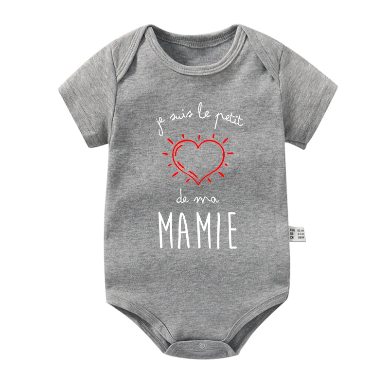 Une combinaison bébé grise à manches courtes, confectionnée en doux Body Coton Manches Courtes de BABY-PREMA, comporte le texte "Je suis le petit coeur de ma mamie" en blanc et rouge, entourant un cœur rouge avec des lignes qui en rayonnent. Parfaite comme cadeau pour bébé, la combinaison est dotée d'une fermeture à bouton-pression en bas et d'une petite étiquette sur la couture du côté gauche.