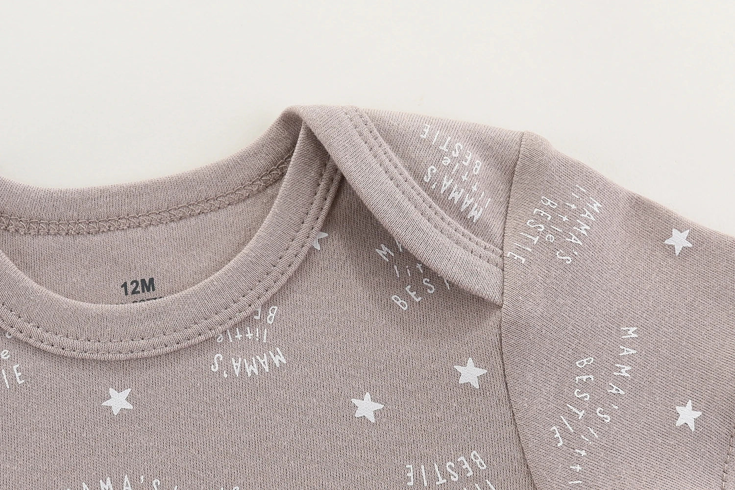 Gros plan d'une combinaison grise pour bébé avec des motifs d'étoiles et de texte blancs, dont « MAMA'S BESTIE ». Cet adorable Lot 6 Body Coton Bébé Manches Courtes de BABY PREMA présente un décolleté enveloppe et une étiquette de taille visible indiquant 12M. Parfait comme cadeau de naissance, il est confectionné en coton avec des manches courtes pour plus de confort.