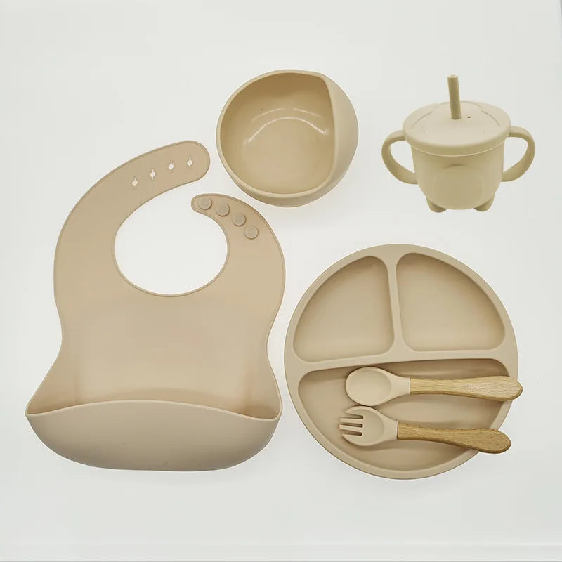 Un ensemble d'alimentation pour bébé beige, l'Ensemble 6-8 Pièces de Vaisselle pour enfants de BABY PREMA, fabriqué en silicone de haute qualité, comprenant un bavoir, un bol, un gobelet, une assiette divisée, une cuillère et une fourchette disposés sur un fond blanc.