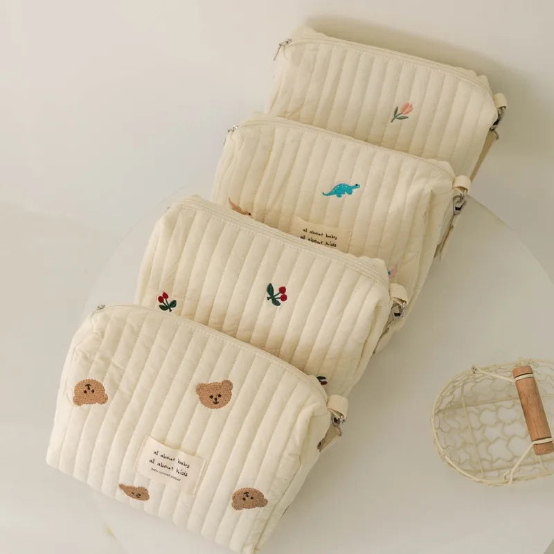 Un lot de quatre pochettes BABY PREMA Sac à Couches en Coton pour poussette en tissu matelassé de couleur crème avec de jolies broderies d'animaux et de fleurs, disposées en cascade sur un fond clair. Ces pochettes poids léger sont parfaites pour voyager.