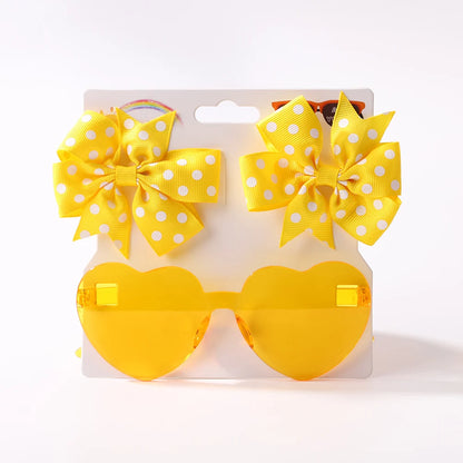 Des lunettes de soleil jaunes en forme de cœur associées à deux nœuds pour cheveux jaunes à pois blancs, présentés sur un fond uni : idéal Set d'Accessoires Bébé | Lunettes de Cheveux pour Bébé de BABY PREMA.