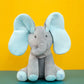 Un tout doux BABY-PREMA Doudou Eléphant Peluche Musicale avec des oreilles bleues et une écharpe à pois posée sur un fond jaune et vert, parfait pour bébé.