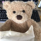 Un ours en peluche Peekaboo moelleux qui sort d'un sac en tissu de couleur crème sur un bureau avec un ordinateur en arrière-plan, prêt pour le dodo de Baby Prema.
