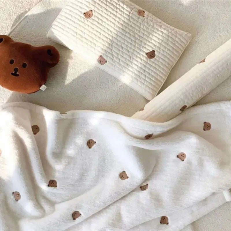 Un arrangement douillet composé d'une Couverture de Bébé en Molleton de Corail de BABY PREMA, d'un coussin tête d'ours en peluche ludique, d'un tissu blanc plié orné de jolis motifs d'ours et d'un coussin blanc immaculé, le tout disposé de manière harmonieuse.