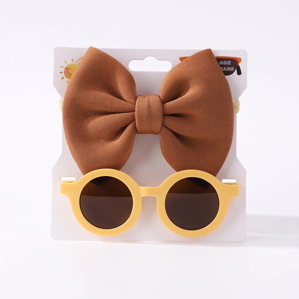 Une paire de lunettes de soleil rondes jaunes pour bébé avec des verres foncés et un grand bandeau à nœud marron, affichées sur un fond blanc. L'Ensemble d'Accessoires Bébé | Lunettes de Cheveux pour Bébé de BABY PREMA.