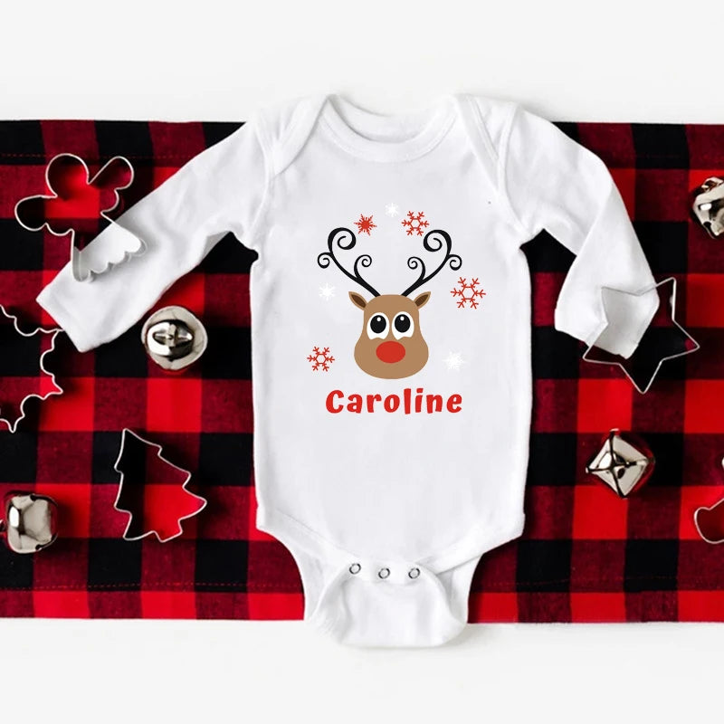 Une combinaison blanche pour bébé avec un visage de renne avec des bois, orné de flocons de neige rouges et du nom « Caroline » en dessous. Posé sur un tissu à carreaux rouges et noirs, ce Body Noël Personnalisé pour Bébé de BABY-PREMA est entouré d'objets festifs comme des cloches et des emporte-pièces en forme d'arbres et d'étoiles, un parfait exemple de vêtements de fête bébé.