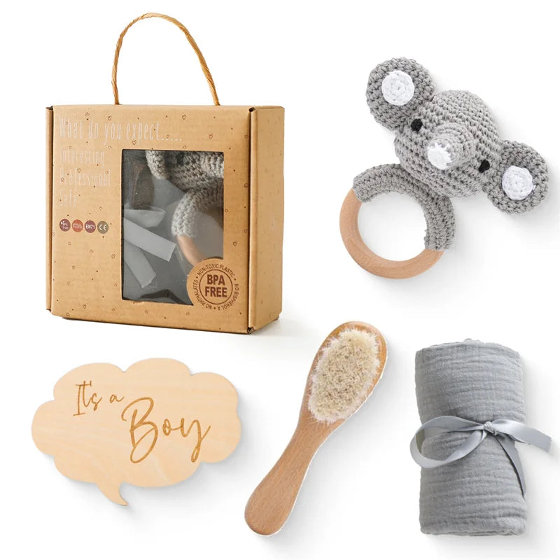 Une collection d'articles BABY PREMA Coffret Cadeau Naissance Bébé comprenant un jouet éléphant au crochet, une brosse en bois à poils naturels, une couverture grise toute douce et un emballage en carton avec fenêtre transparente, le tout disposé sur un fond neutre