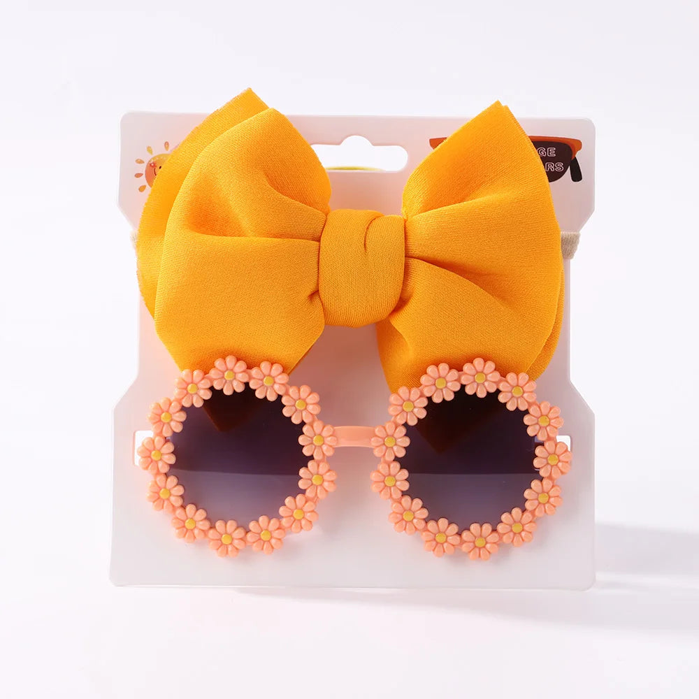 Un paquet contenant une paire de lunettes de soleil rondes avec une monture florale rose et un grand nœud en tissu jaune, des bandeaux à nœuds en nylon exposés sur un fond blanc. 

Phrase corrigée : Un paquet contenant le Set d'Accessoires Bébé | Lunettes de Cheveux pour Bébé de BABY PREMA exposées sur fond blanc.