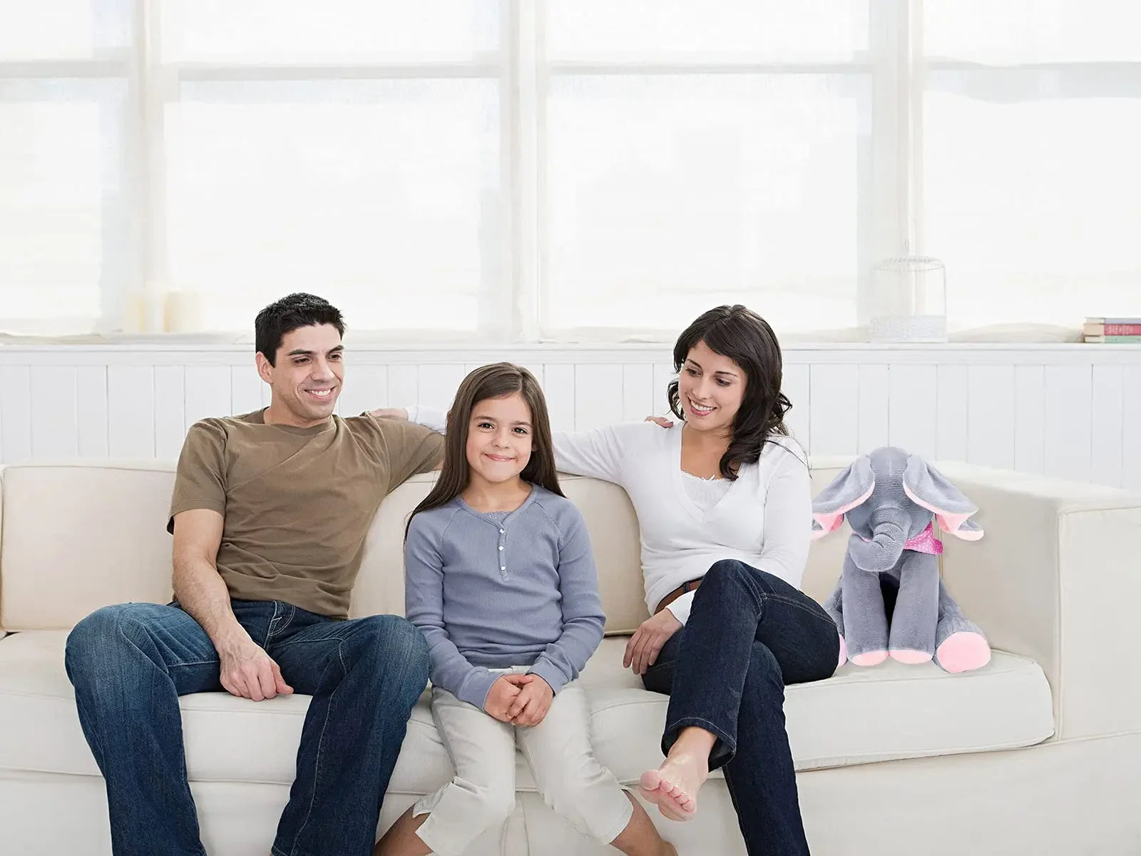 Une famille de trois personnes, composée d'un homme et d'une femme encadrant une jeune fille, confortablement assis ensemble sur un canapé blanc dans une pièce bien éclairée, partageant un moment de convivialité avec le sourire en jouant avec le BABY-PREMA Jouet éléphant en peluche | chantant avec Oreilles Mobiles.