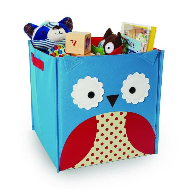 Une boîte de rangement colorée pour enfants conçue pour ressembler à un sympathique hibou, remplie de divers jouets et livres, créant un espace amusant et bien rangé pour les enfants. Cette Caisse Rangement pliable Hiboux de Skip Hop se replie facilement.