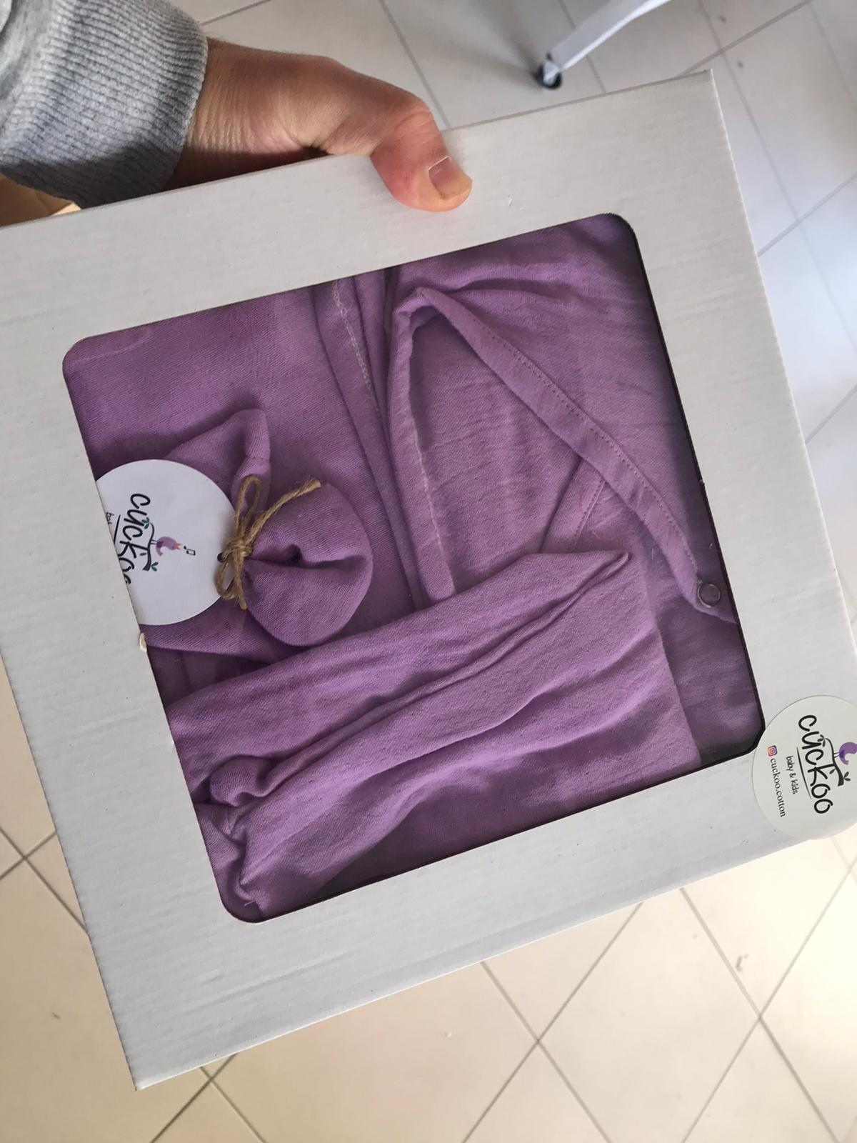 Une personne tenant un ensemble emballé de literie pour bébé violette Carte Cadeaux Naissance avec une étiquette visible de la marque BABY-PREMA.